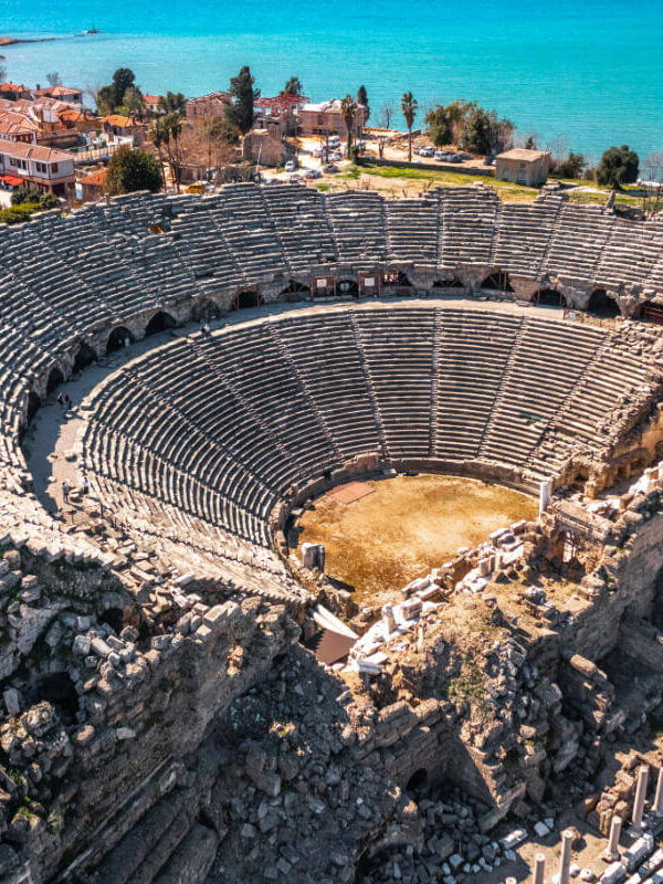 Aerial view of the Ancient Theater of Side in Antalya, Turkey, with stone seating and coastal town in the background.