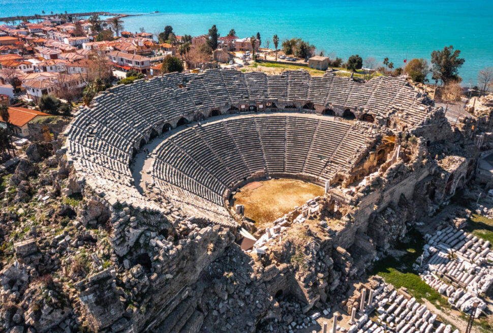 Aerial view of the Ancient Theater of Side in Antalya, Turkey, with stone seating and coastal town in the background.