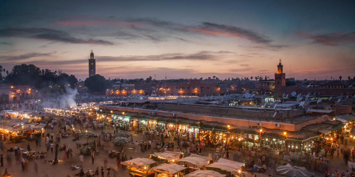 Bustling Djemaa el-Fna square in Marrakech at sunset with food stalls, lights, and silhouettes under colorful sky.