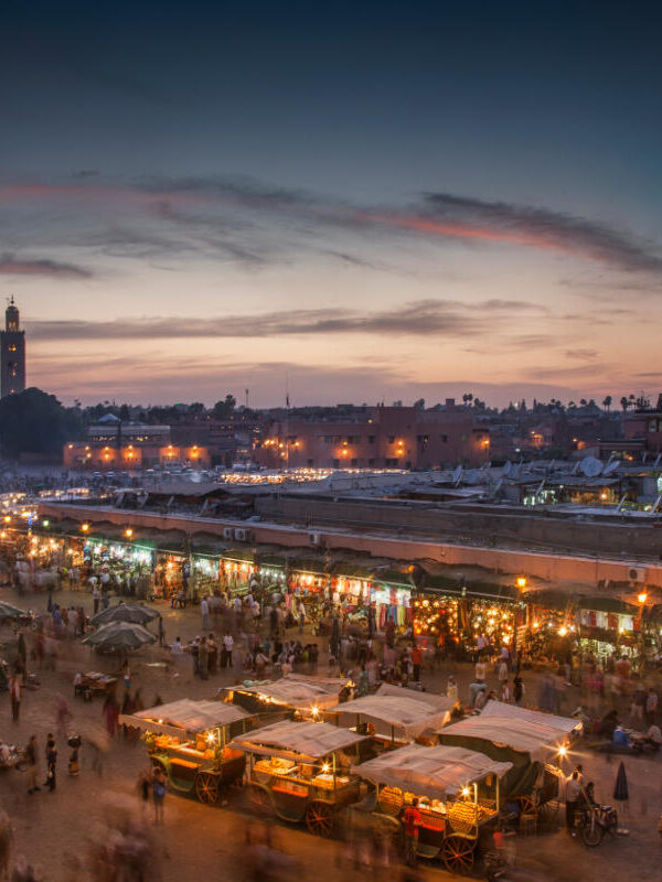 Bustling Djemaa el-Fna square in Marrakech at sunset with food stalls, lights, and silhouettes under colorful sky.