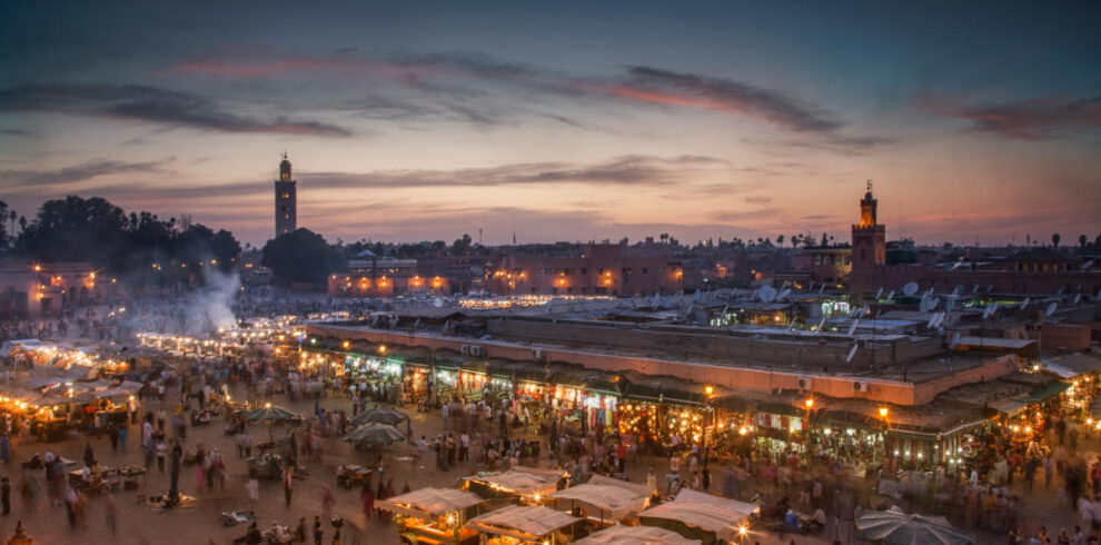 Bustling Djemaa el-Fna square in Marrakech at sunset with food stalls, lights, and silhouettes under colorful sky.