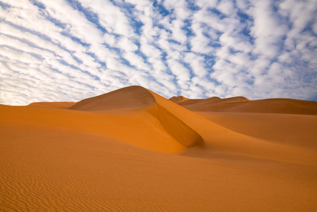 Wavy golden sand dunes in a desert landscape beneath a patterned sky filled with white scattered clouds.