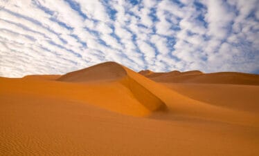 Wavy golden sand dunes in a desert landscape beneath a patterned sky filled with white scattered clouds.