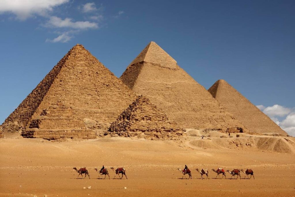 Camel caravan passing in front of Egypt's Giza pyramids under a clear blue sky in desert landscape.