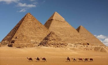Camel caravan passing in front of Egypt's Giza pyramids under a clear blue sky in desert landscape.