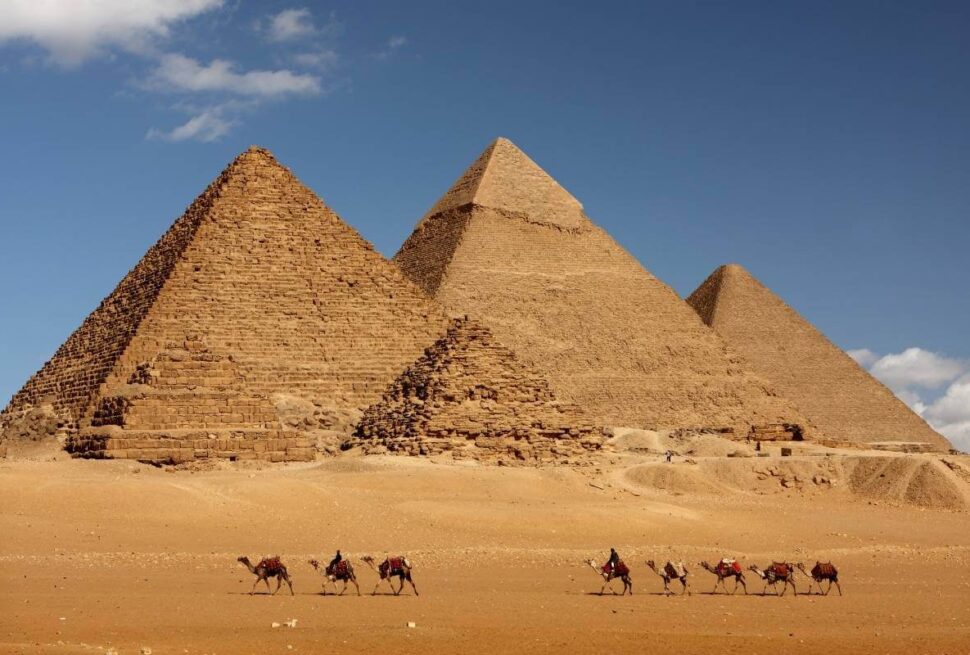 Camel caravan passing in front of Egypt's Giza pyramids under a clear blue sky in desert landscape.