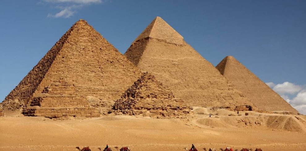 Camel caravan passing in front of Egypt's Giza pyramids under a clear blue sky in desert landscape.