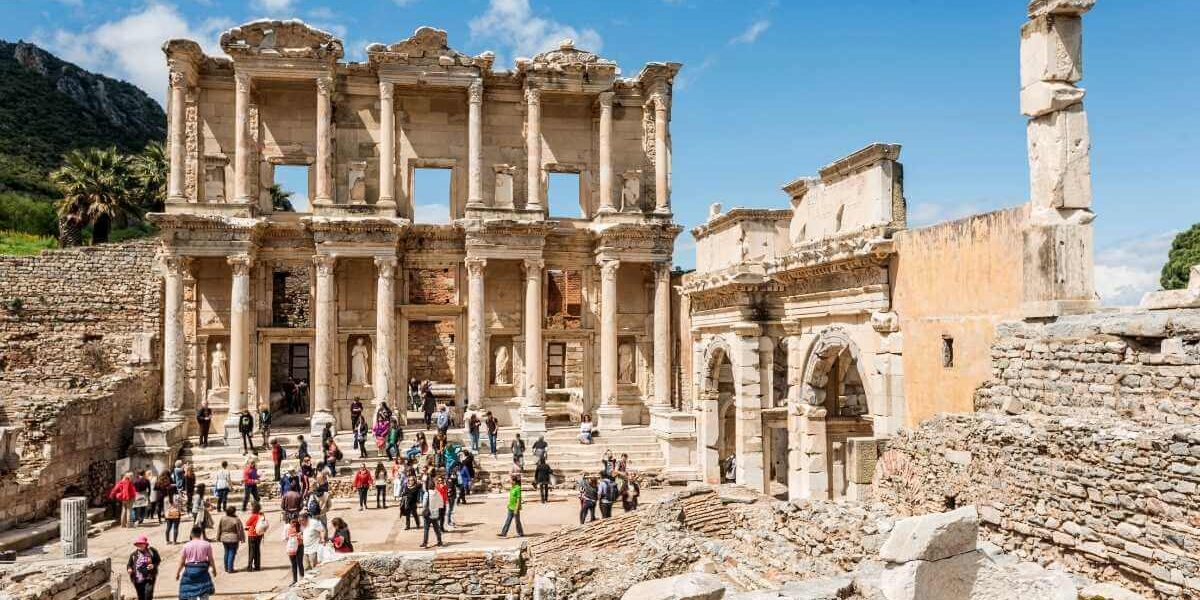 Tourists exploring the grand façade of the ancient Library of Celsus in Ephesus with clear blue skies above.