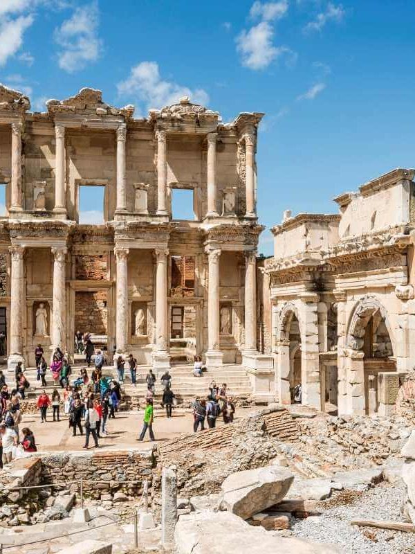 Tourists exploring the grand façade of the ancient Library of Celsus in Ephesus with clear blue skies above.