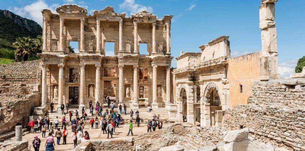Tourists exploring the grand façade of the ancient Library of Celsus in Ephesus with clear blue skies above.