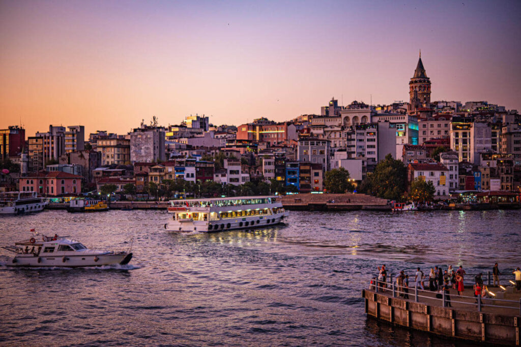 Galata Tower above Istanbul’s skyline at sunset with boats cruising the Golden Horn and people gathered on the waterfront.
