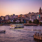 Galata Tower above Istanbul’s skyline at sunset with boats cruising the Golden Horn and people gathered on the waterfront.
