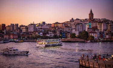 Galata Tower above Istanbul’s skyline at sunset with boats cruising the Golden Horn and people gathered on the waterfront.