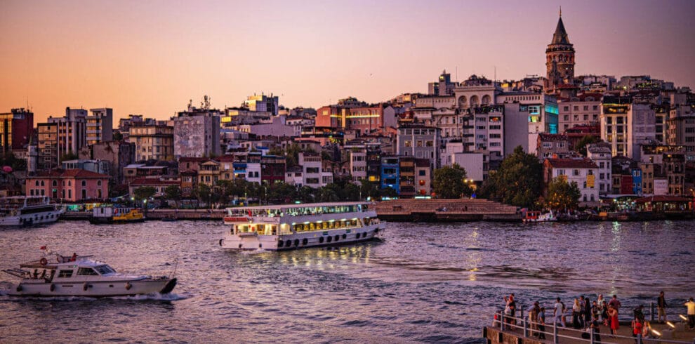 Galata Tower above Istanbul’s skyline at sunset with boats cruising the Golden Horn and people gathered on the waterfront.