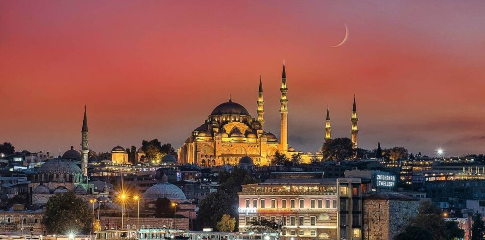 Illuminated Suleymaniye Mosque in Istanbul at sunset with boats on the Bosphorus and a crescent moon above.