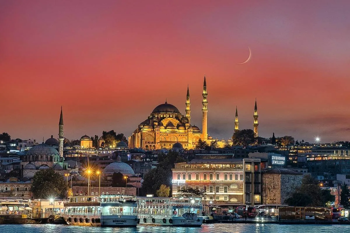 Illuminated Suleymaniye Mosque in Istanbul at sunset with boats on the Bosphorus and a crescent moon above.