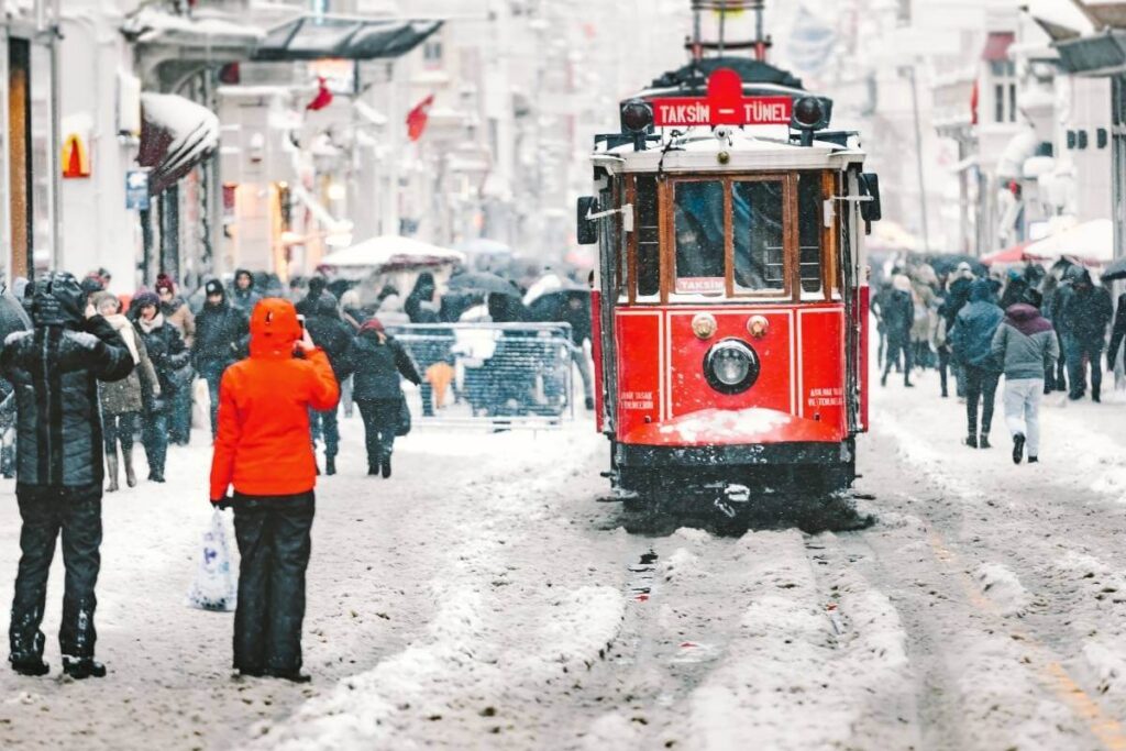 Iconic red Taksim tram moves through snow-covered Istiklal Street surrounded by winter-clad pedestrians in Istanbul.