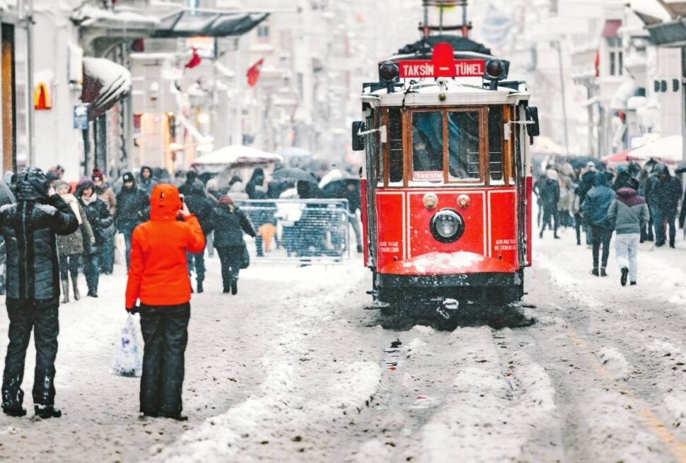Iconic red Taksim tram moves through snow-covered Istiklal Street surrounded by winter-clad pedestrians in Istanbul.