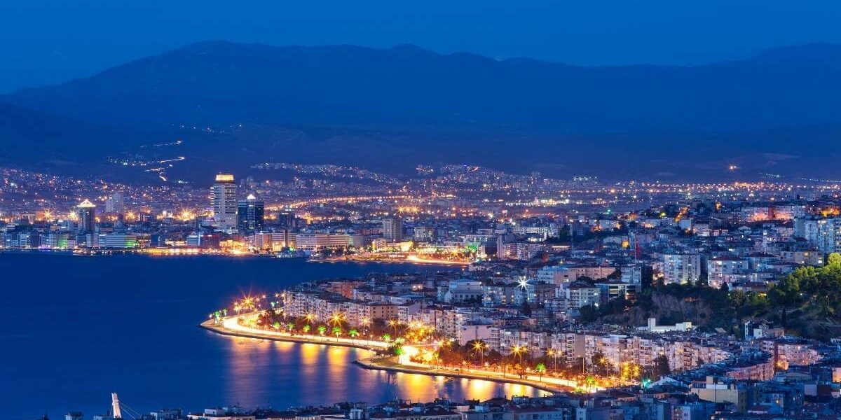 Aerial night view of İzmir's illuminated waterfront, showcasing coastal buildings and city lights by the bay.