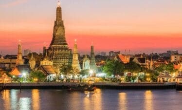 Wat Arun temple illuminated at sunset along the Chao Phraya River in Bangkok, Thailand.
