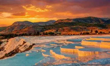 A vibrant sunset casts golden and orange light over the white travertine thermal pools of Pamukkale, Turkey, with tiered turquoise water basins filling the landscape against rugged mountains.