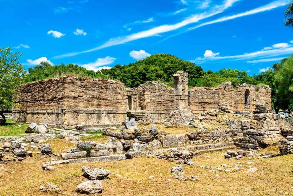 Stone ruins of an ancient structure in Olympia, Greece, surrounded by trees and a bright blue summer sky.