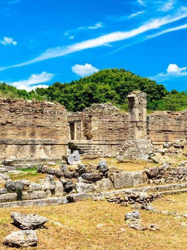 Stone ruins of an ancient structure in Olympia, Greece, surrounded by trees and a bright blue summer sky.
