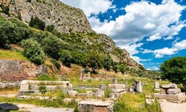 Stone ruins of Ancient Delphi in Greece surrounded by green hills and mountains under a bright blue sky.