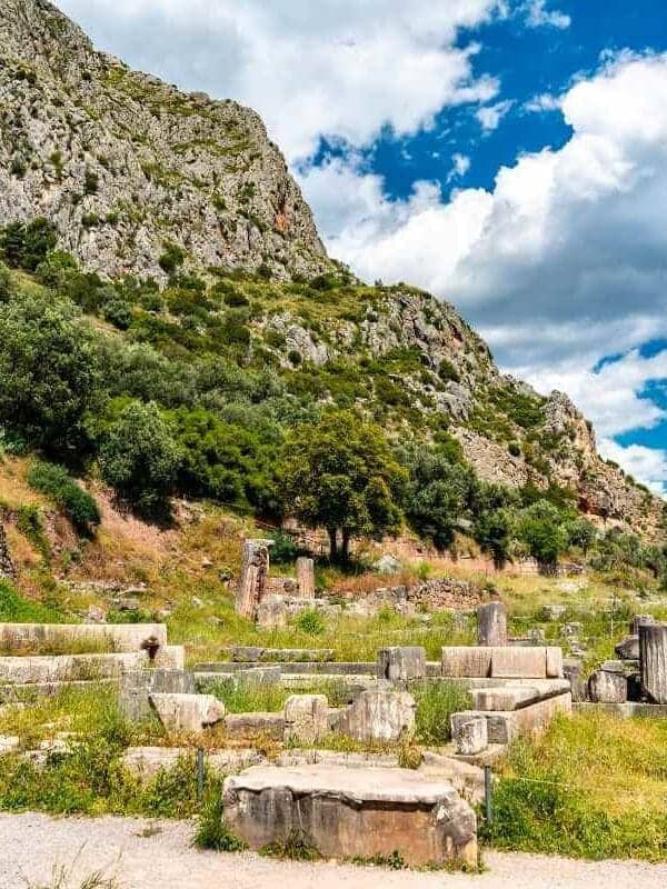 Ruinas de piedra de la antigua Delfos en Grecia, rodeadas de verdes colinas y montañas bajo un cielo azul brillante.