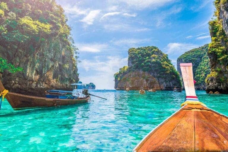 Traditional Thai longtail boats float on turquoise waters near limestone cliffs in a tropical Thailand bay.