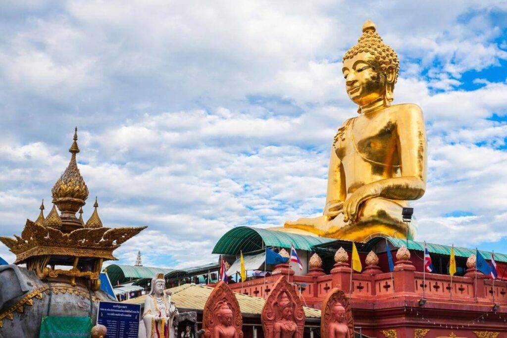 Massive golden Buddha statue atop red temple base with flags and ornate statues in Chiang Rai, Thailand.