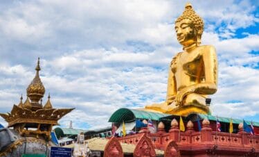 Massive golden Buddha statue atop red temple base with flags and ornate statues in Chiang Rai, Thailand.