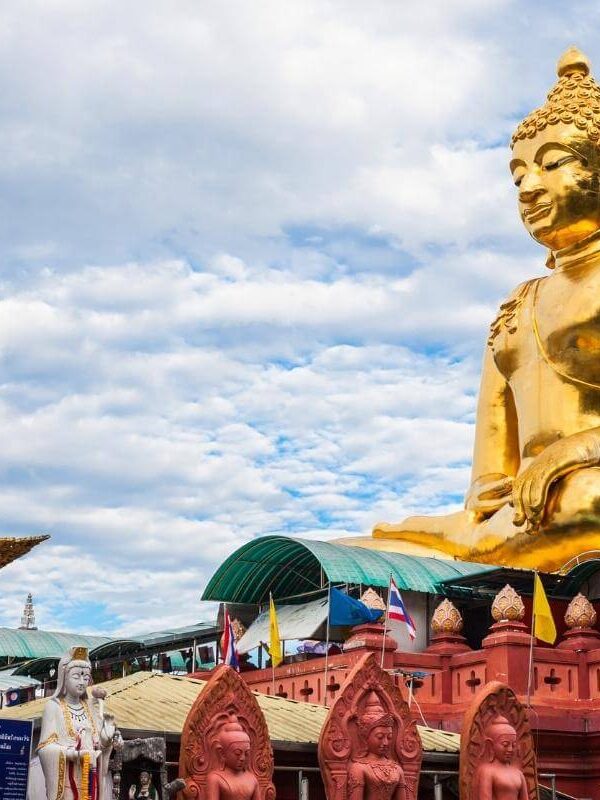 Massive golden Buddha statue atop red temple base with flags and ornate statues in Chiang Rai, Thailand.