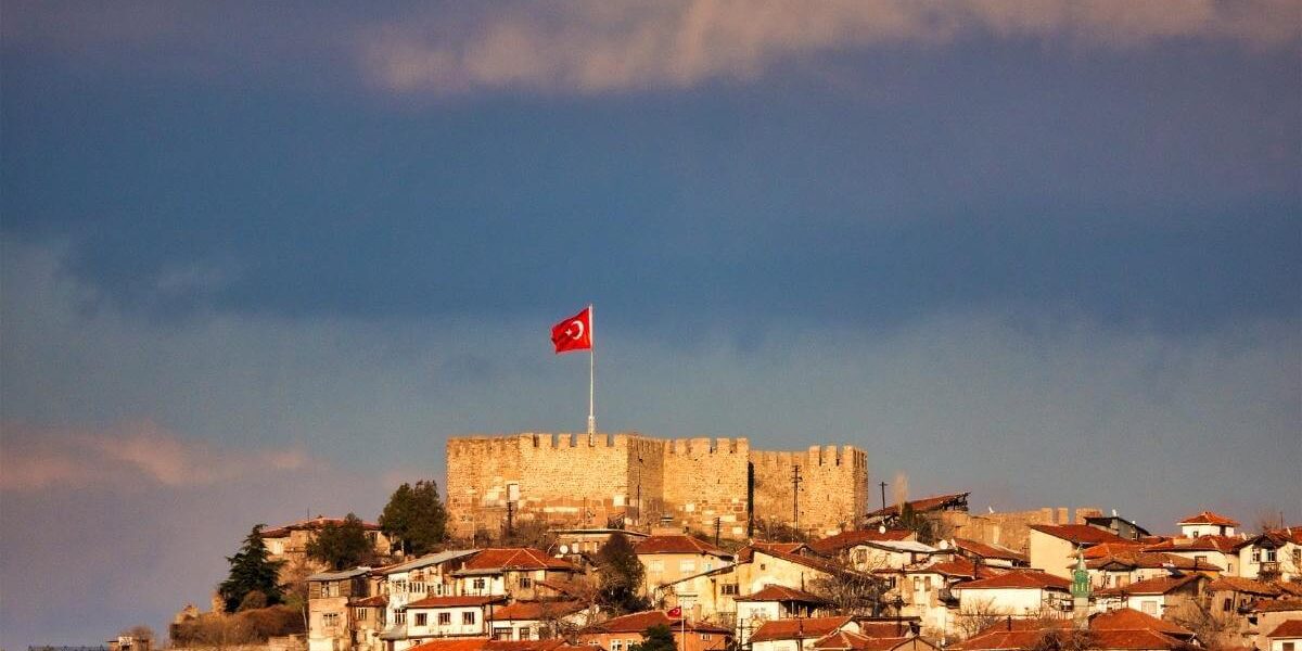 Historic Ankara Castle with Turkish flag overlooking old town houses under dramatic sky.