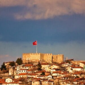 Historic Ankara Castle with Turkish flag overlooking old town houses under dramatic sky.