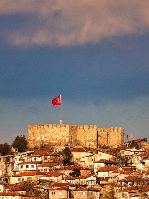 Historic Ankara Castle with Turkish flag overlooking old town houses under dramatic sky.