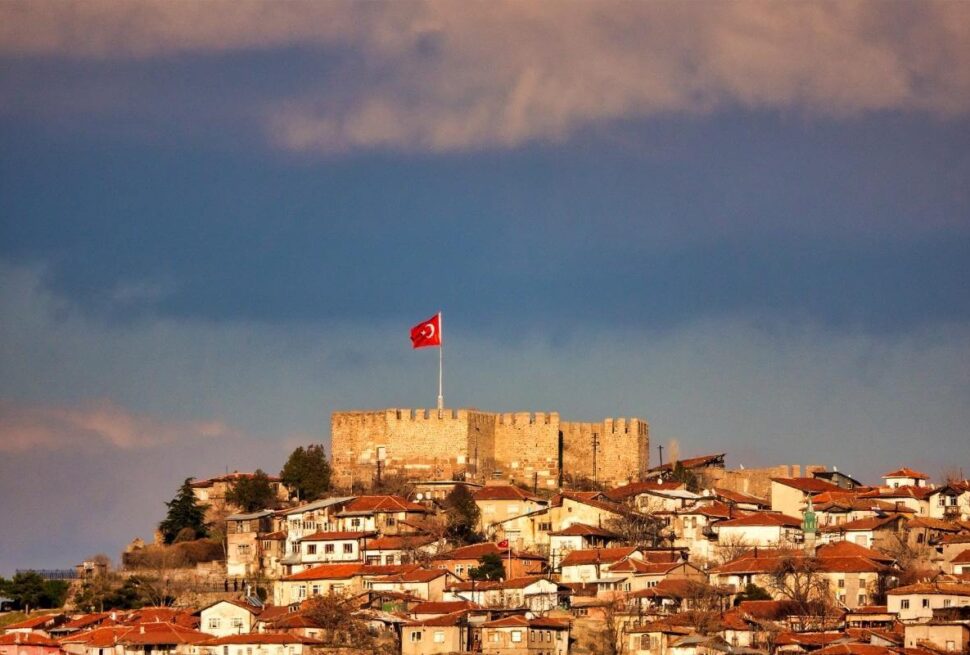 Historic Ankara Castle with Turkish flag overlooking old town houses under dramatic sky.