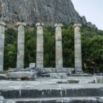 Ancient stone columns of the Temple of Athena in Priene, Turkey, with a mountain backdrop and lush green trees.