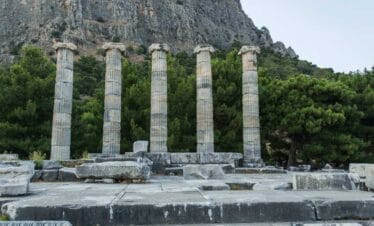 Ancient stone columns of the Temple of Athena in Priene, Turkey, with a mountain backdrop and lush green trees.