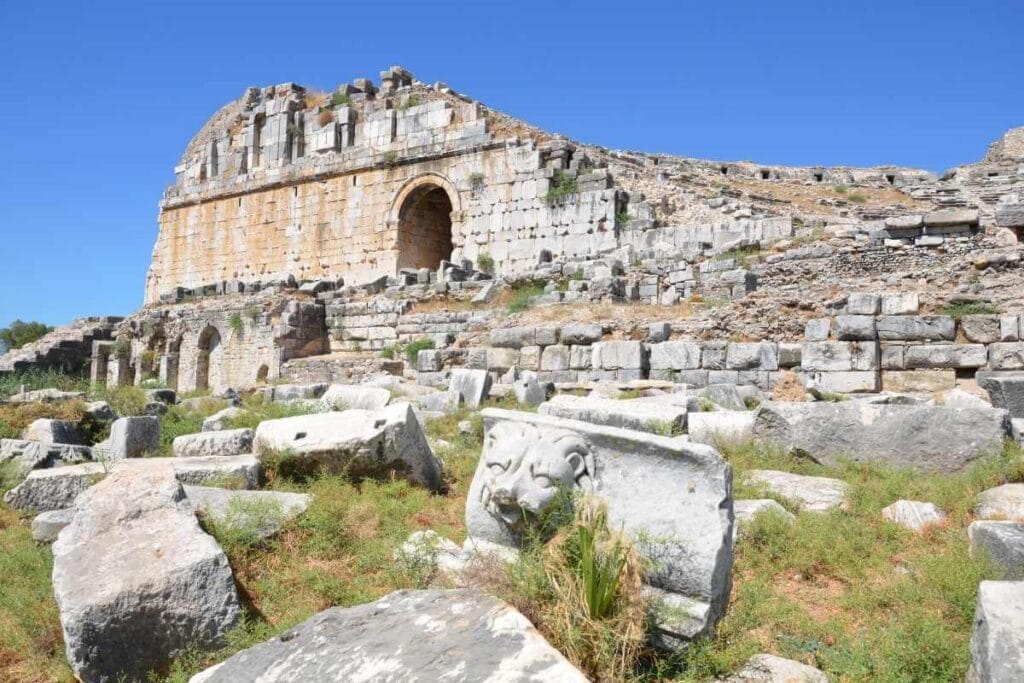 Ruins of Miletos Ancient Theatre in Turkey with stone seating, arched entrance, and carved lion relief under a clear blue sky.