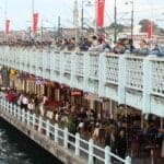 Crowds fishing on Galata Bridge in Istanbul above waterfront restaurants with Turkish flags and city skyline views.