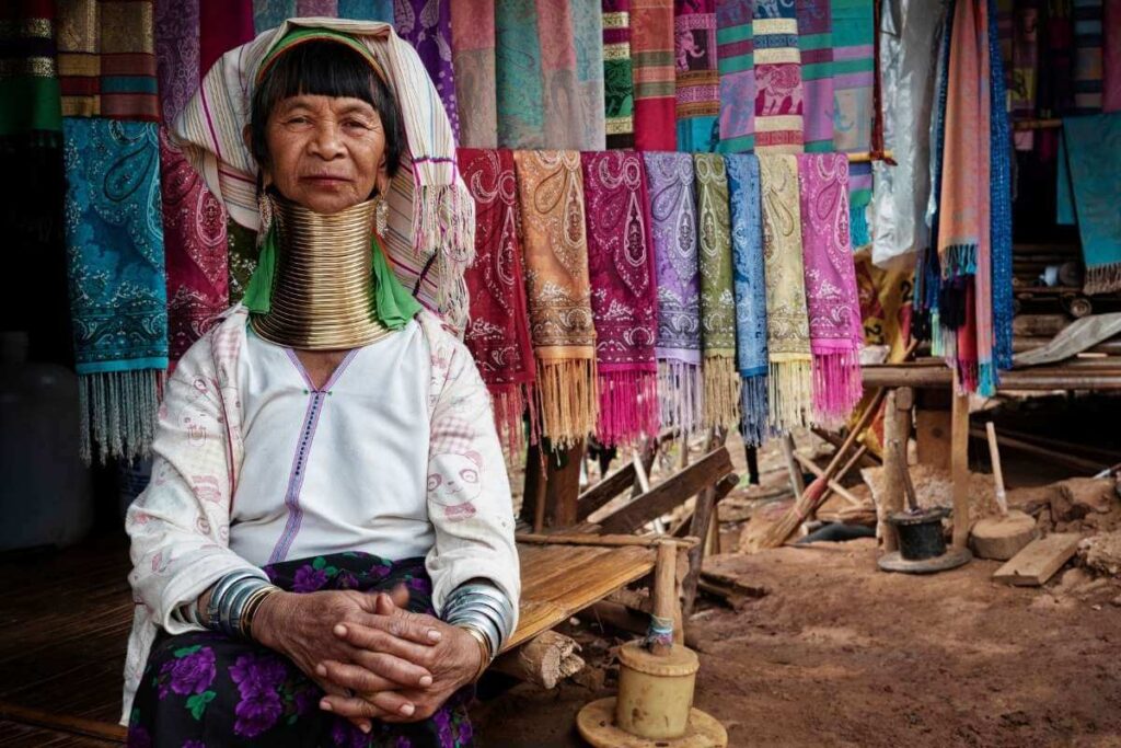 Karen Long Neck woman in traditional attire with brass neck rings, sitting beside colorful handmade scarves in Thailand.