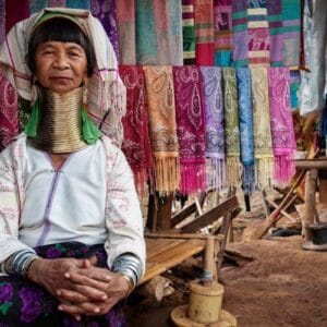 Karen Long Neck woman in traditional attire with brass neck rings, sitting beside colorful handmade scarves in Thailand.
