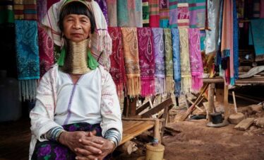 Karen Long Neck woman in traditional attire with brass neck rings, sitting beside colorful handmade scarves in Thailand.