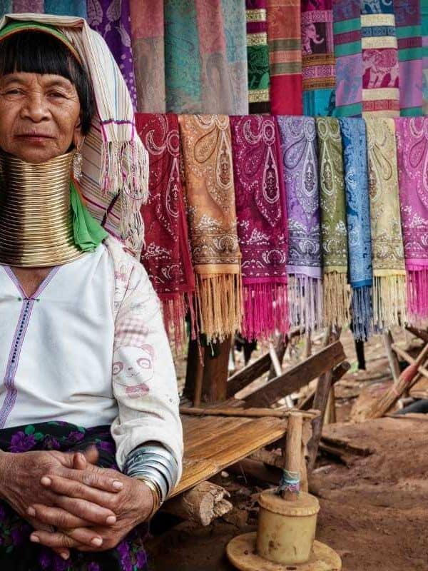 Karen Long Neck woman in traditional attire with brass neck rings, sitting beside colorful handmade scarves in Thailand.