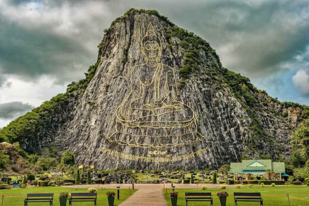 Giant Buddha image engraved in gold on Khao Chi Chan mountain cliff in Pattaya, Thailand, with lush greenery below.