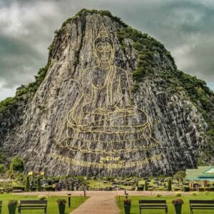 Giant Buddha image engraved in gold on Khao Chi Chan mountain cliff in Pattaya, Thailand, with lush greenery below.