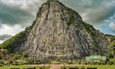 Giant Buddha image engraved in gold on Khao Chi Chan mountain cliff in Pattaya, Thailand, with lush greenery below.
