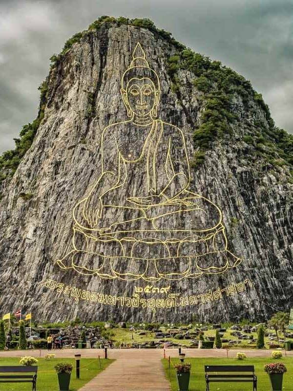 Giant Buddha image engraved in gold on Khao Chi Chan mountain cliff in Pattaya, Thailand, with lush greenery below.