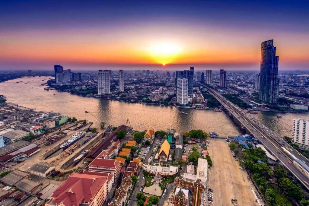 Aerial view of Bangkok city skyline with Chao Phraya River at sunset, featuring skyscrapers, bridges, and colorful temples.